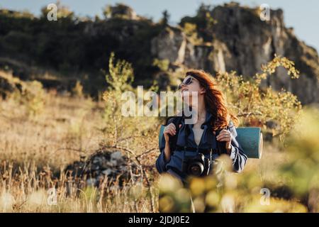 Junge Reisende Frau in Sonnenbrillen mit Digitalkamera und Rucksack mit Campingmatte die Wanderin steht gegen den Berg und genießt Freiheit auf ihrem Solo Stockfoto