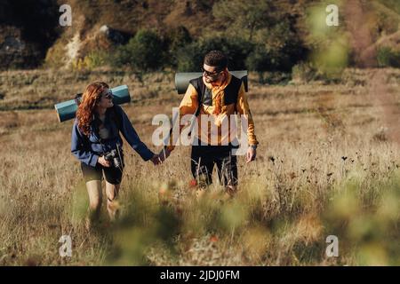 Mann und Frau halten sich bei Sonnenuntergang an den Händen, während sie gemeinsam auf dem Hügel wandern, und das junge Paar genießt ihre Reise mit Rucksäcken und Campingmatte Stockfoto