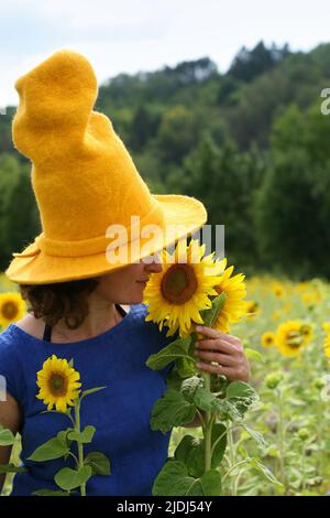 Frau mit braunem kurzen lockigen Haar, das königsblaues Kleid trägt und einen großen gelben Hut mit einem breiten Rand, der nach Sonnenblume riecht. Wandern im Sonnenblumenfeld Stockfoto