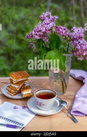 Eine Teeparty mit Wiener Waffeln und einem Zweig Flieder und einem offenen leeren Notizbuch zum Schreiben auf einem Holztisch vor dem Hintergrund eines Gartens Stockfoto