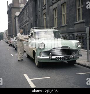 1964, historisch, steht ein Mann in seiner Arbeit vor seinem Auto der Zeit, ein Ton-Ton, Ford Zodiac, geparkt in einer Straße, England, Großbritannien Stockfoto