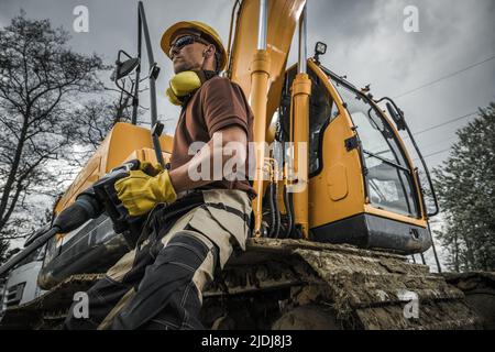 Kaukasischer Bauarbeiter in seinem 40s stolzen Halten eines Presshammers vor dem Big Bagger Digger. Heavy Duty Maschinen Thema. Stockfoto