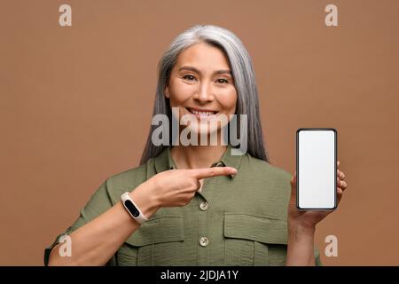 Porträt einer älteren älteren lächelnden Frau, die eine Handyanwendung zeigt oder vorstellt und mit dem Finger auf den Smartphone-Bildschirm zeigt, während eine reife Geschäftsfrau auf braunem Hintergrund isoliert ist Stockfoto