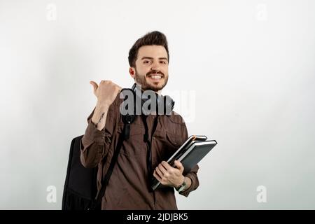 Junger Mann mit braunem Hemd, der isoliert auf weißem Hintergrund posiert und lächelt, mit einem glücklichen Gesicht, das mit dem Daumen nach oben zur Seite zeigt. Stockfoto