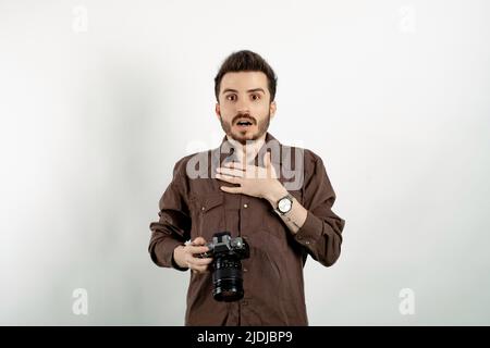 Hübscher junger Mann in legerer Kleidung, isoliert auf weißem Hintergrund, schockiert und überrascht, lächelnd und während des Urlaubs Hand an Herz nehmend Stockfoto