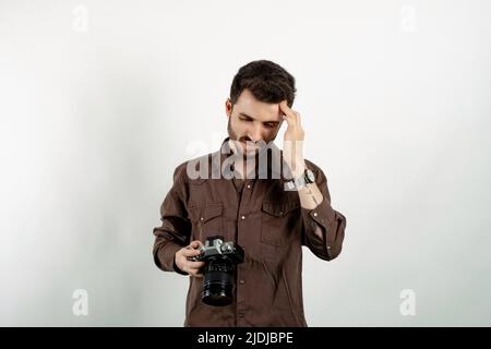 Kaukasischer Mann mit braunem Hemd, der isoliert auf weißem Hintergrund posiert, gestresst, unglücklich und frustriert fühlt, die Stirn berührt und dslr C hält Stockfoto