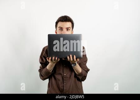 Junger Mann in braunem Hemd, der isoliert auf weißem Hintergrund posiert und hinter dem Laptop aus der Kamera schaut. Stockfoto