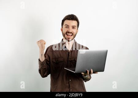 Fröhlicher kaukasischer Mann in braunem Hemd, der isoliert auf weißem Hintergrund mit einem Laptop-Computer posiert und dabei eine Gewinnergeste macht. Stockfoto