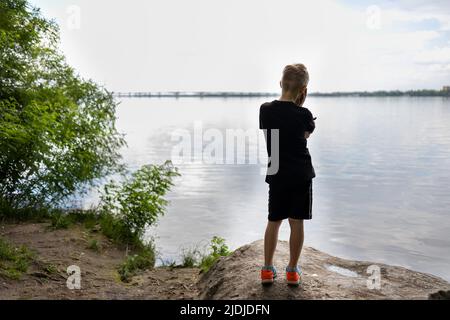 Der junge Reisende steht mit dem Rücken auf einem Stein und blickt nachdenklich auf den Fluss Stockfoto