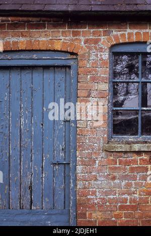 Detail der alten orangen Backstein stabilen Block oder Scheune mit abblätternden blau lackierten Holztür und Fenstern und Schiefer Dach Stockfoto