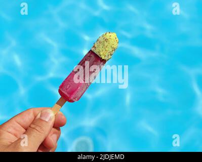 Die Hand hält fruchtige bunte Eiszapfen oder Wassereis vor einer blauen Pooloberfläche, helfen gegen Sommerhitze Konzeptbild Stockfoto