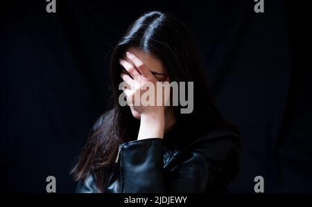 Junge Frau in schwarzer Lederjacke, die Augen, Scham und Bedauern Gefühle in einem dunklen Raum bedeckt. Angst und Verzweiflung Konzept Stockfoto
