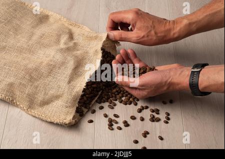 Kaffeebohnen, die in der Hand liegen, werden aus einer hessischen Tasche auf hellem Holzhintergrund aus nächster Nähe verschüttet Stockfoto