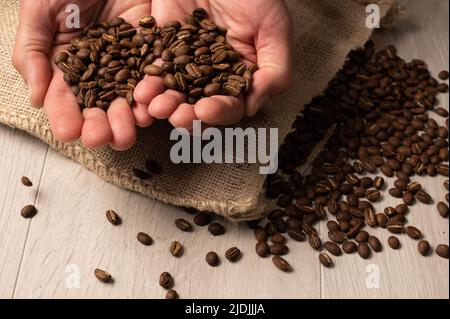 Kaffeebohnen, die in der Hand liegen, werden aus einer hessischen Tasche auf hellem Holzhintergrund aus nächster Nähe verschüttet Stockfoto