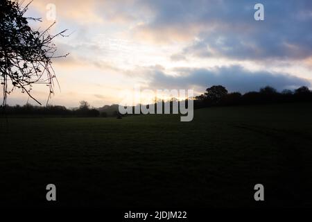 Gelber Sonnenuntergang über den Hügeln und Bäumen und grünen Feldern mit Sturmwolken und einem hellblauen Himmel Stockfoto