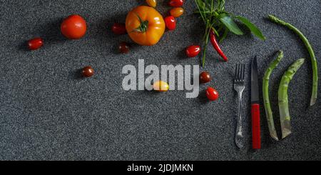 Lebensmittelrand mit grünem Spargel, roten und orangen Tomaten und rotem Chili mit Blättern. Gemüse, rotes Messer und Gabel auf dunkelgrauem Hintergrund t Stockfoto