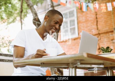 Lächelnder und zufriedener multirassischer Geschäftsmann arbeitet online mit einem Laptop und trinkt Kaffee im Freien. Freiberufliche Außenstelle Stockfoto
