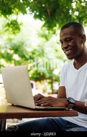 Nahaufnahme eines lachenden Geschäftsmannes mit gemischter Rasse, der mit einem Laptop arbeitet und als freiberuflicher Technologiearbeiter im Straßencafé online arbeitet. Stockfoto