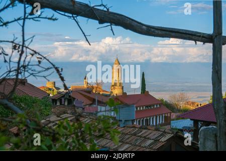 Sonniger Kirchturm und rote Dächer von Häusern, die sich hinter Baumzweigen gegen den wolkigen blauen Himmel auf der Straße von Sighnaghi, Georgia, befinden Stockfoto