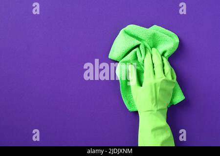 Weibliche Hand in grünem Schutzhandschuh mit grünem Lappen auf violettem Hintergrund. Housekeeping Service und Housekeeping Konzept. Stockfoto
