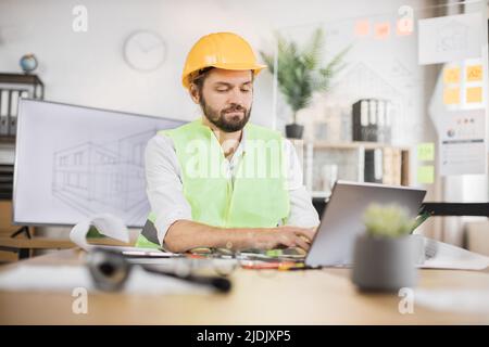 Kompetenter Ingenieur, Designer oder Architekt, der die Arbeitskonferenz im Bürozentrum leitet. Attraktiver junger bärtiger Mann mit Uniform und Schutzhelm, der am Laptop mit Blaupause arbeitet. Stockfoto