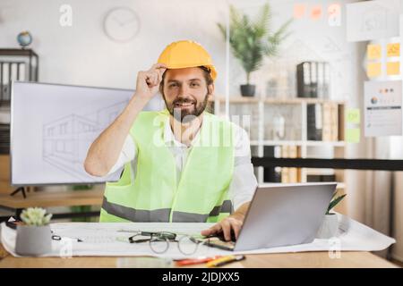 Junger Architekt männlich in Helm und Uniform sitzen am Tisch mit Blaupausen und modernen Geräten. Fokussierter kaukasischer Arbeiter bei der Erstellung eines neuen Bauprojekts, der in einem modernen Büro sitzt. Stockfoto