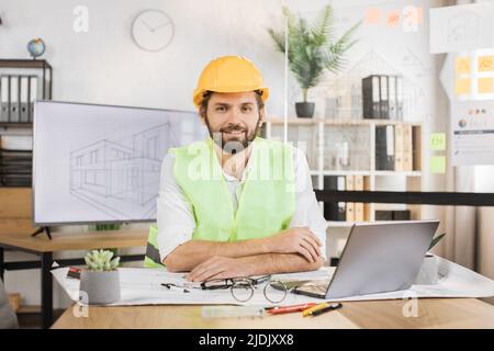 Kompetenter männlicher Architekt, der im Innenbereich mit einem digitalen Laptop auf dem Schreibtisch sitzt und den Kompass für Projektzeichnungen verwendet. Positiv bärtiger junger Mann mit einheitlicher grüner reflektierender Weste und einem harten Hut aus der Konstruktion. Stockfoto