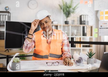 Attraktiver afrikanischer Ingenieur in weißem Helm und reflektierender Weste, der am Tisch sitzt, mit vielen Blaupausen auf dem Hintergrund eines großen fernsehbildschirms mit Skizze. Geschäftsmann, der an einem Bauprojekt arbeitet. Stockfoto