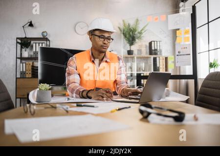 Junger Architekt afrikanischer Mann in Helm und Uniform sitzt am Tisch mit Blaupausen und modernen Geräten. Fokussierter Arbeiter bei der Erstellung eines neuen Bauprojekts, der in einem modernen Büro sitzt. Stockfoto