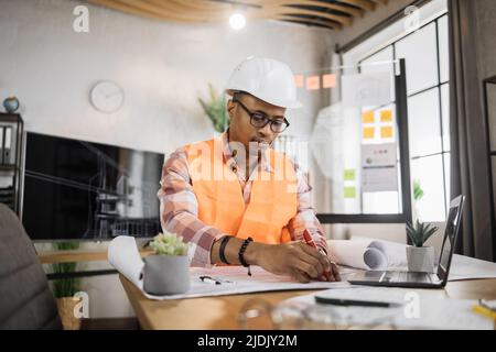 Kompetenter afroamerikanischer Ingenieur, Designer oder Architekt, der die Arbeitskonferenz im Bürozentrum leitet. Attraktiver junger Mann mit Uniform und Schutzhelm, der am Laptop mit Blaupause arbeitet. Stockfoto