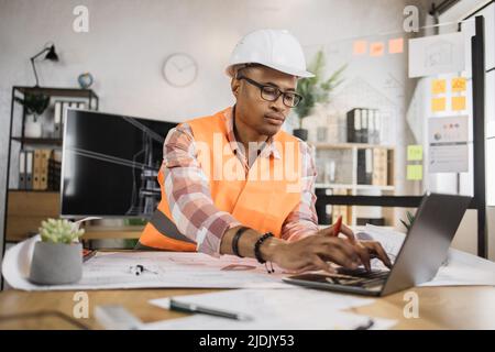 Junger Architekt afrikanischer Mann in Helm und Uniform sitzt am Tisch mit Blaupausen und modernen Geräten. Fokussierter Arbeiter bei der Erstellung eines neuen Bauprojekts, der in einem modernen Büro sitzt. Stockfoto