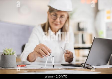Mit dem Kompass auf die Hand fokussieren. Leitende Ingenieurin Frau in weißem Helm und formeller Kleidung sitzt am Tisch mit vielen Blaupausen auf dem Hintergrund eines großen fernsehbildschirms mit Skizze. Stockfoto