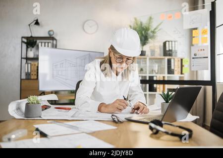 Kompetente, reife Ingenieurin, Designerin oder Architektin, die die Arbeitskonferenz im Bürozentrum leitet. Attraktive junge ältere Frau mit Schutzhelm, die mit Kompass, Laptop und Blaupause arbeitet. Stockfoto