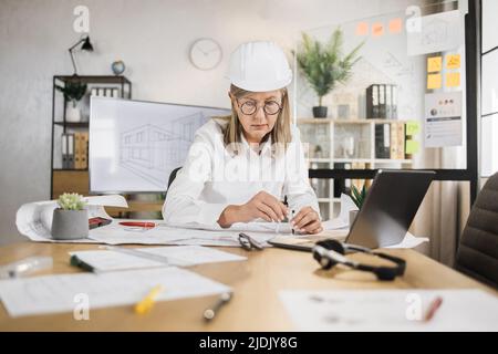 Kompetente, reife Ingenieurin, Designerin oder Architektin, die die Arbeitskonferenz im Bürozentrum leitet. Attraktive junge ältere Frau mit Schutzhelm, die mit Kompass, Laptop und Blaupause arbeitet. Stockfoto