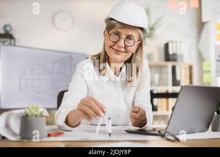 Kompetente, reife Ingenieurin, Designerin oder Architektin, die die Arbeitskonferenz im Bürozentrum leitet. Attraktive junge ältere Frau mit Schutzhelm, die mit Kompass, Laptop und Blaupause arbeitet. Stockfoto