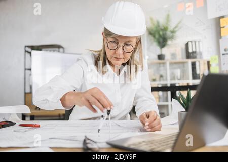 Kompetente, reife Ingenieurin, Designerin oder Architektin, die die Arbeitskonferenz im Bürozentrum leitet. Attraktive junge ältere Frau mit Schutzhelm, die mit Kompass, Laptop und Blaupause arbeitet. Stockfoto