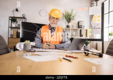 Kompetenter, reifer Ingenieur, Designer oder Architekt, der die Arbeitskonferenz im Bürozentrum leitet. Attraktiver älterer bärtiger Mann mit Uniform und Schutzhelm, der auf einem Laptop mit Blaupause arbeitet. Stockfoto