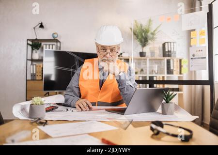 Kompetenter, reifer Ingenieur, Designer oder Architekt, der die Arbeitskonferenz im Bürozentrum leitet. Attraktiver älterer bärtiger Mann mit Uniform und Schutzhelm, der auf einem Laptop mit Blaupause arbeitet. Stockfoto