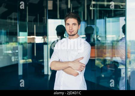 Porträt eines hübschen jungen männlichen IT-Spezialisten, Freiberufler. In weißen Kleidern im Büro stehend, Arme gekreuzt, lächelnd auf die Kamera schauend Stockfoto