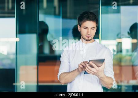 Porträt eines jungen, gutaussehenden männlichen Geschäftsmannes, Freiberufler. Im Büro stehend und ein Tablet in den Händen haltend, arbeitend, tippend. Stockfoto