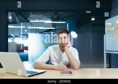 Langweiliger Arbeitstag. Porträt eines jungen Mannes, der traurig im Büro am Schreibtisch sitzt und seinen Kopf in der Hand hält. Er mag seinen Job nicht. Müde Stockfoto