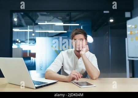 Langweiliger Arbeitstag. Porträt eines jungen Mannes, der traurig im Büro am Schreibtisch sitzt und seinen Kopf in der Hand hält. Er mag seinen Job nicht. Müde Stockfoto