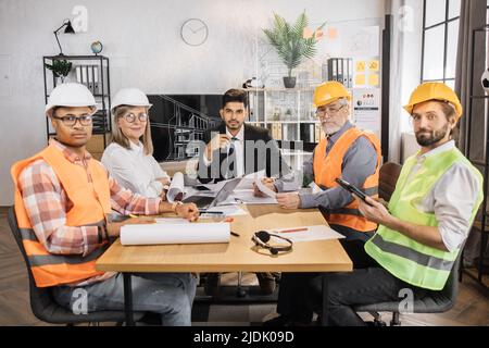 Qualifizierte Architekten, Ingenieure und Designer treffen sich im Büro vor dem Fernsehbildschirm. Multikulturelle Kollegen in Anzügen und Helmen sitzen am Tisch und blicken in die Kamera. Stockfoto