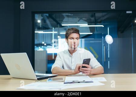 Junger Mann, Geschäftsmann, Manager, der im Büro an seinem Schreibtisch sitzt, ein Mobiltelefon hält, Nachrichten tippt, Anrufe macht. Funktioniert auf Laptops und d Stockfoto