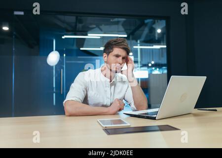 Sich unwohl fühlen. Junger Mann, Freiberufler, Regisseur, Manager, der seinen Kopf hält, Kopfschmerzen hat, müde, krank. Sitzen im Büro an einem Schreibtisch mit einem Lapto Stockfoto