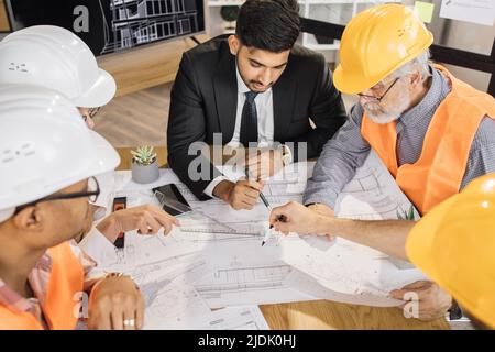 Qualifizierte Architekten, Ingenieure und Designer mit Geschäftstreffen im Büro. Multikulturelle Kollegen in Anzügen und Helmen sitzen am Tisch und schauen sich Blaupausen an. Konstruktionskonzept. Stockfoto