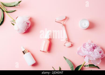 Tropfflaschen mit Kosmetiköl, Serum, Gesichtswalze, Cremeglas und Pfingstrosen auf rosa Hintergrund. Draufsicht, flach liegend. Stockfoto