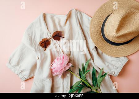 Sommerlicher Casual-Style. Stilvolle Frauenkleidung und Accessoires. Leinenkleid, Sonnenbrille, Hut und Pfingstrose auf rosa Hintergrund. Flach liegend, Draufsicht. Stockfoto