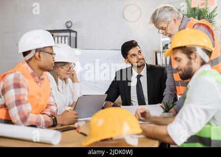 Konzentrieren Sie sich auf schützenden Hardhut. Kompetente Ingenieure, Designer und Architekten leiten die Arbeitskonferenz im Bürozentrum. Gruppe multikultureller Menschen, die Anzug, Uniform und Schutzhelm tragen. Stockfoto