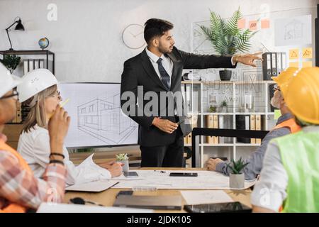 Eine Gruppe von fünf multirassischen Menschen, die formelle Kleidung und Helme tragen und ein gemeinsames Projekt im Büro erstellen und planen. Partner, die in Innenräumen mit Skizzen arbeiten. Stockfoto